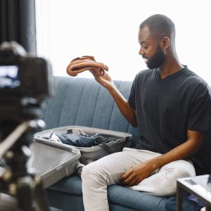 African-american male blogger sitting in front of camera and recording a video about his luggage. Man wearing dark t-shirt and white trousers. Blogger speaking about traveling.